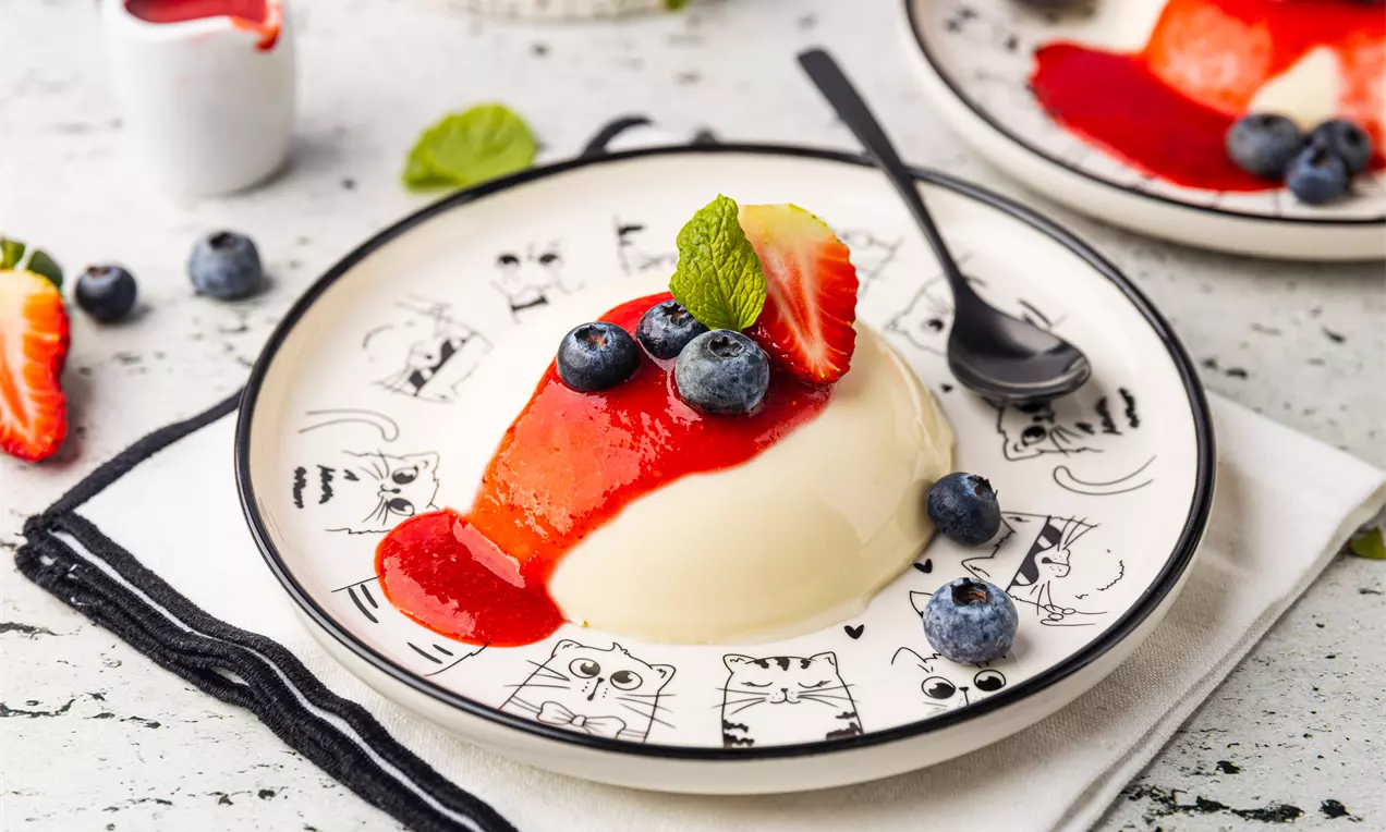 Panna Cotta w kremowej formie z musem truskawkowym, udekorowana borówkami, truskawką i listkiem mięty na talerzu w koty