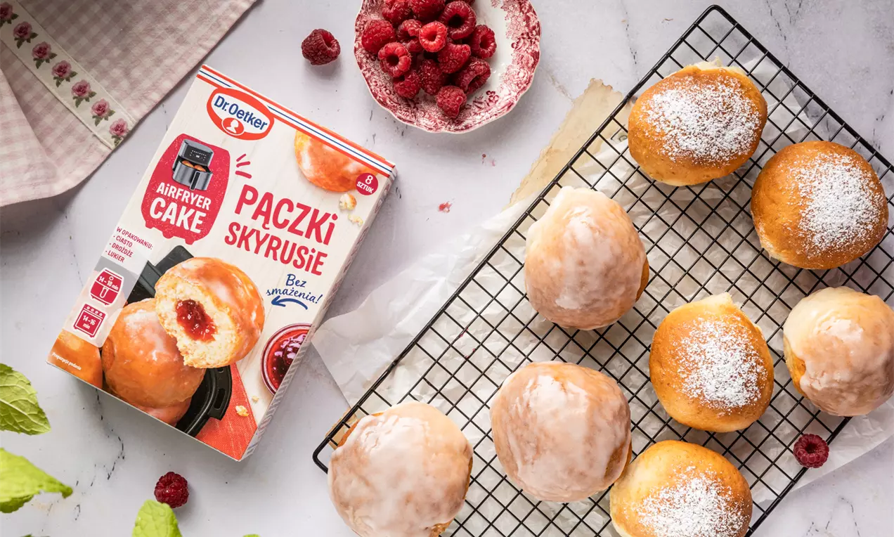 Złociste pączki z lukrem i cukrem pudrem na kratce, obok miseczka z malinami i zielone liście na jasnym tle