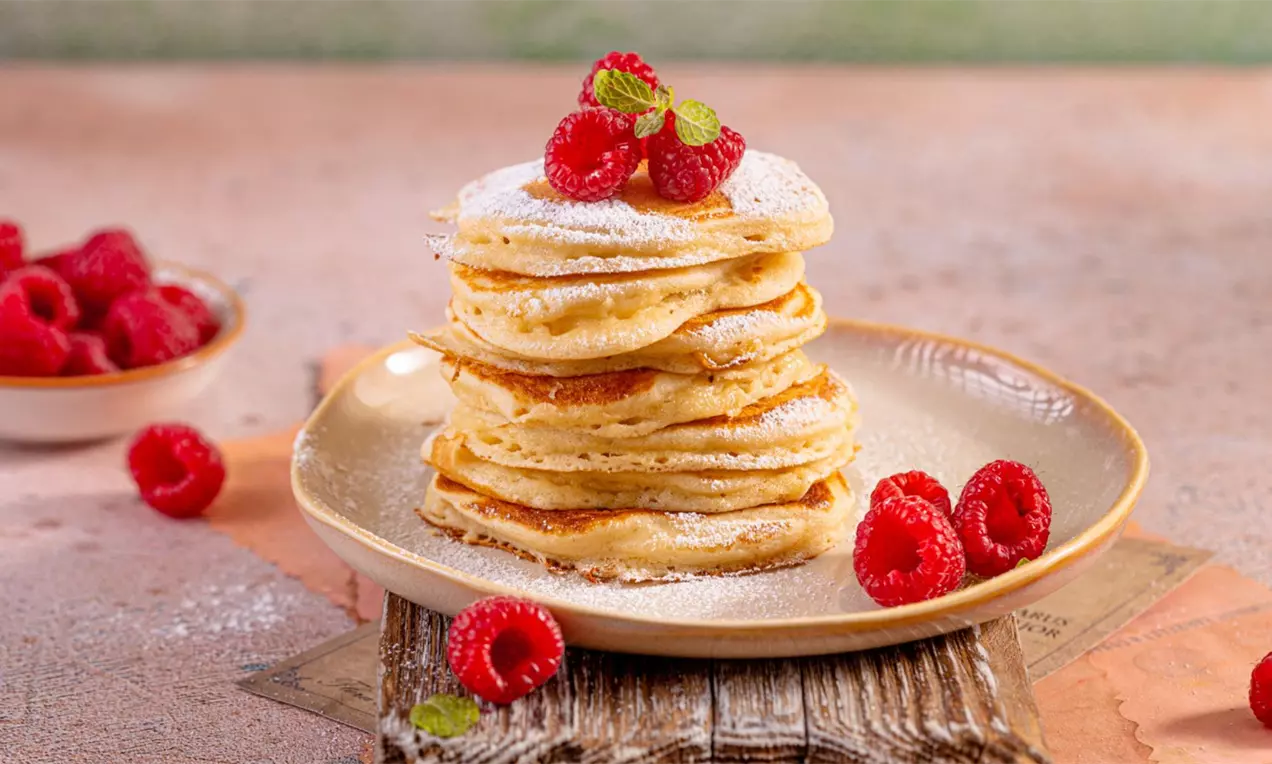Puszyste, złociste pancakes ułożone w stos na jasnym talerzu, udekorowane malinami, cukrem pudrem i listkami mięty