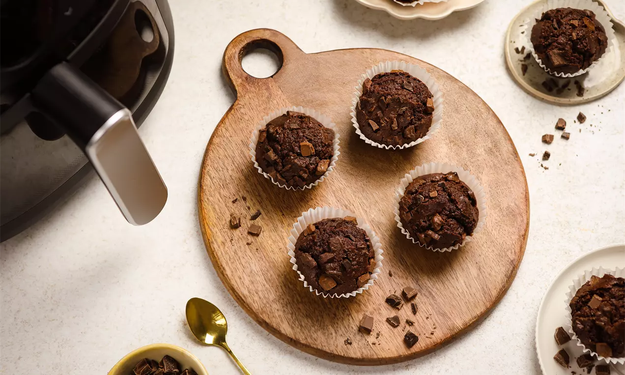 Czekoladowe muffinki w papierowych foremkach, ułożone na okrągłej drewnianej desce, obok kawałków czekolady i złotej łyżeczki