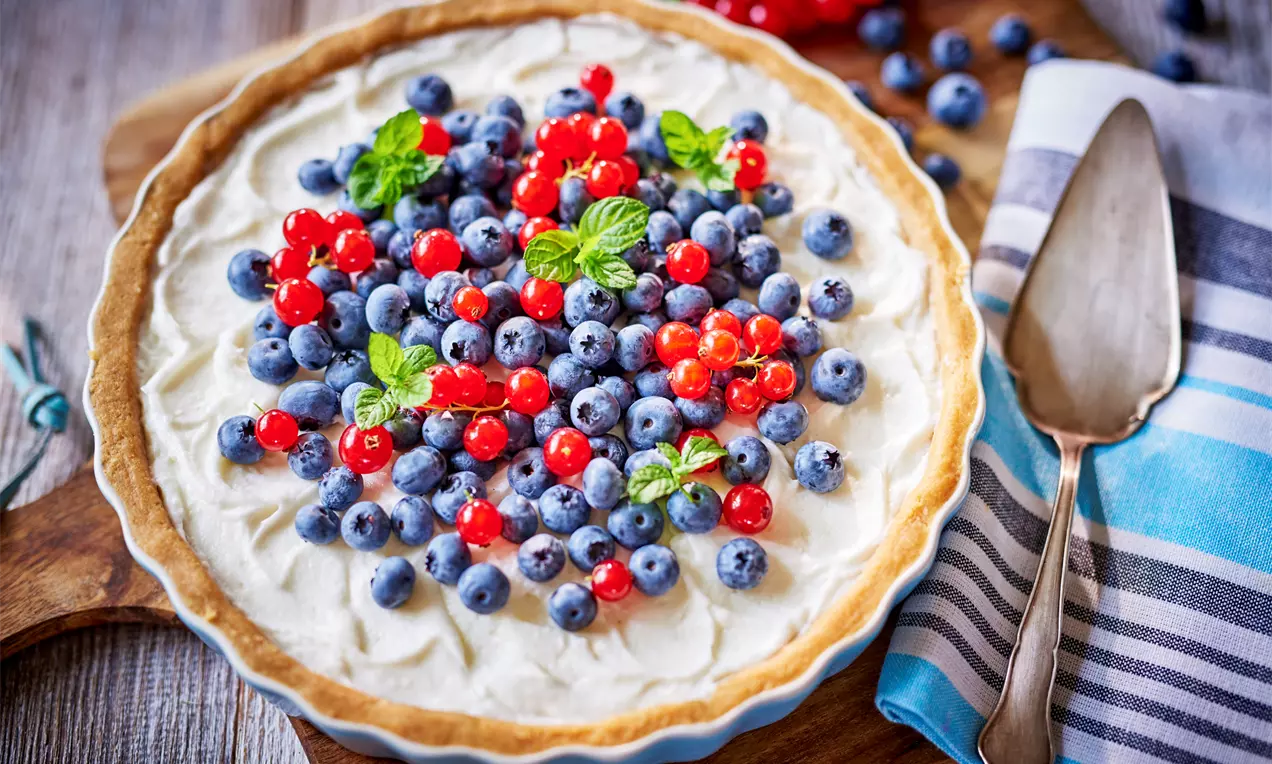 Okrągła tarta na jasnoniebieskiej formie z kremem jogurtowym, udekorowana borówkami, porzeczkami i listkami mięty na drewnianej desce