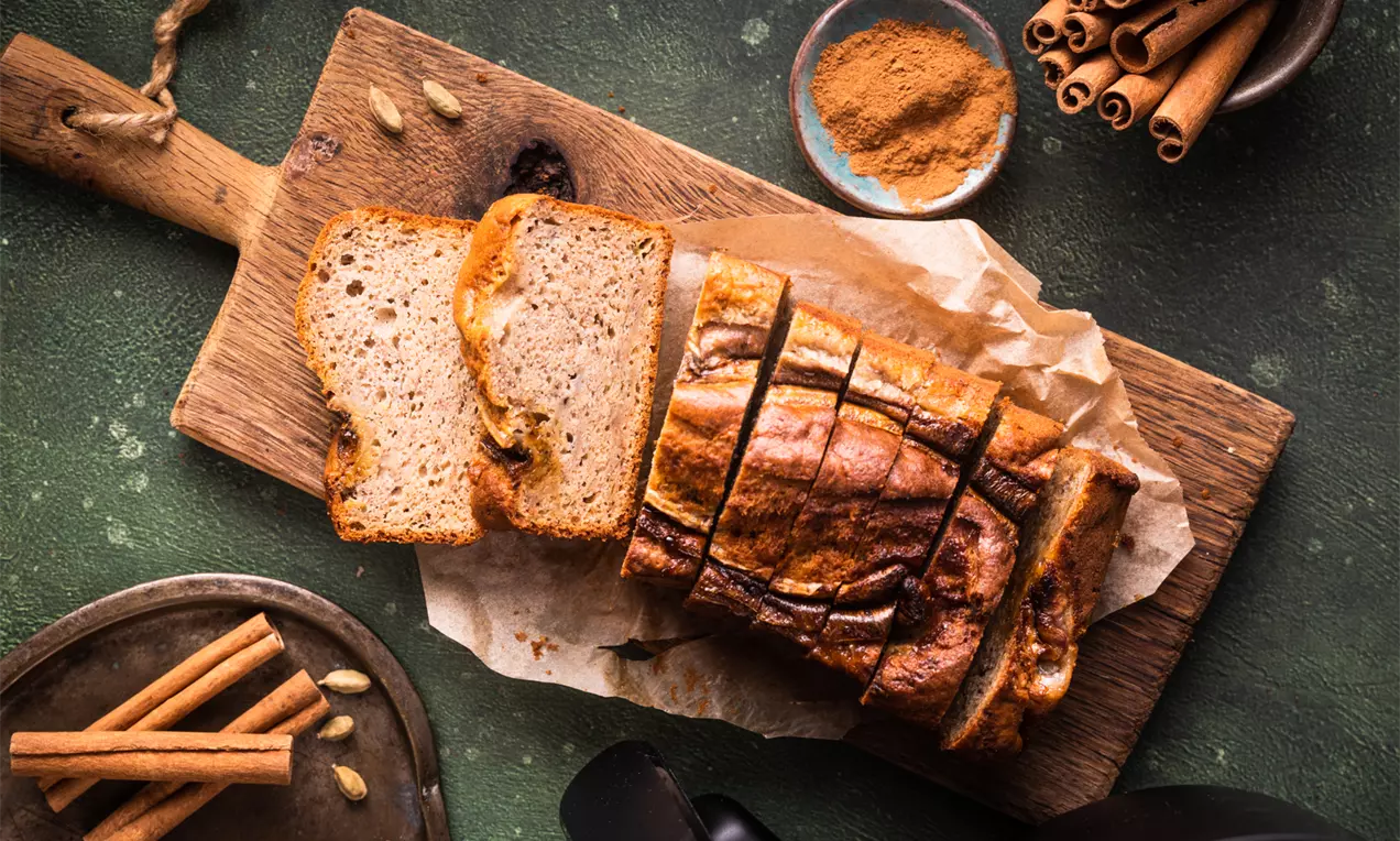 Chlebek bananowy w złocistobrązowym odcieniu, pokrojony na grube kromki, podany na drewnianej desce z pergaminem, obok laski cynamonu i miseczka z przyprawą